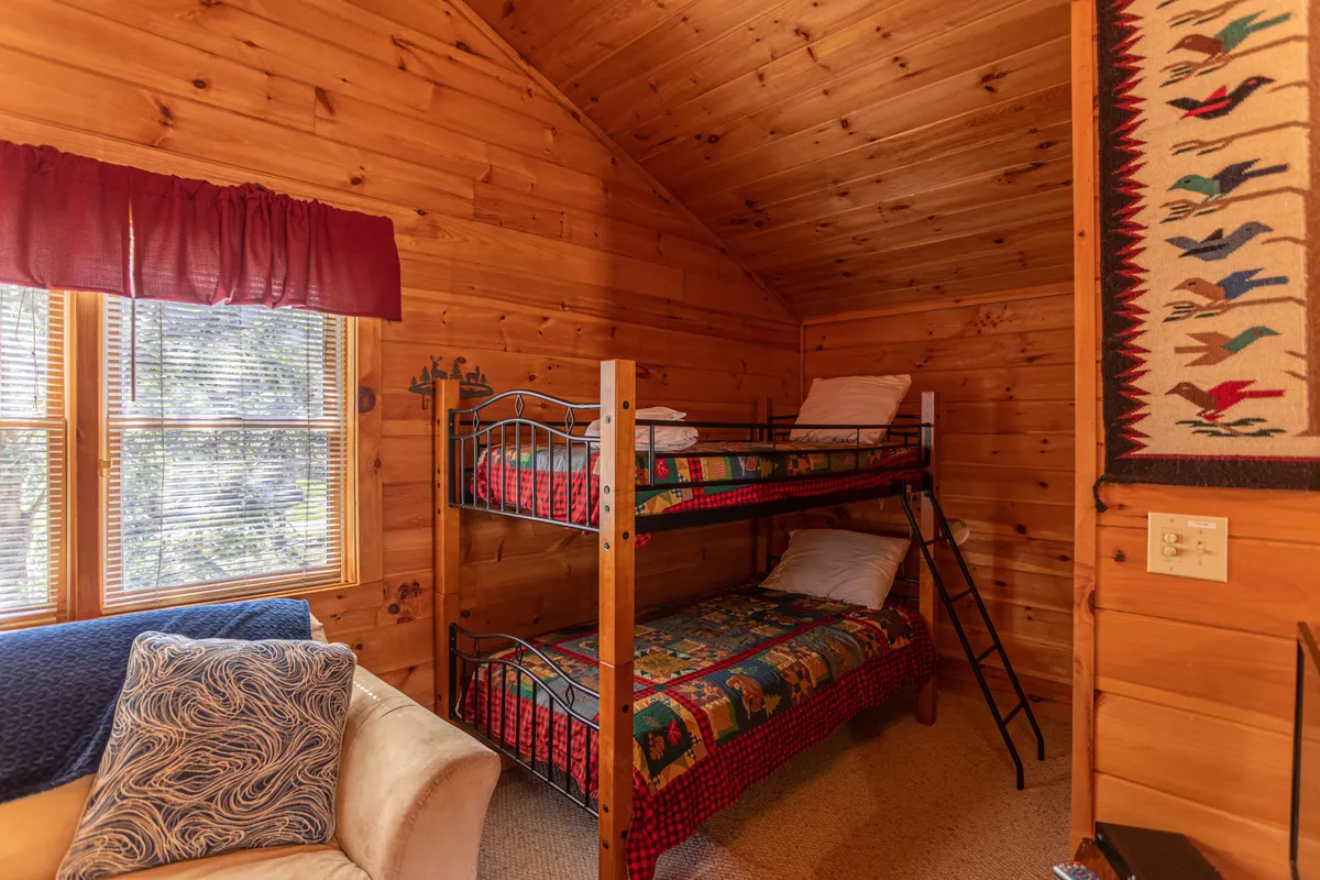 A loft area with twin bunk beds, a sofa nearby, a decorative wall hanging, and a window letting in morning sunbeams. The layout feels youthful, flexible, and perfect for kids or friends to share.