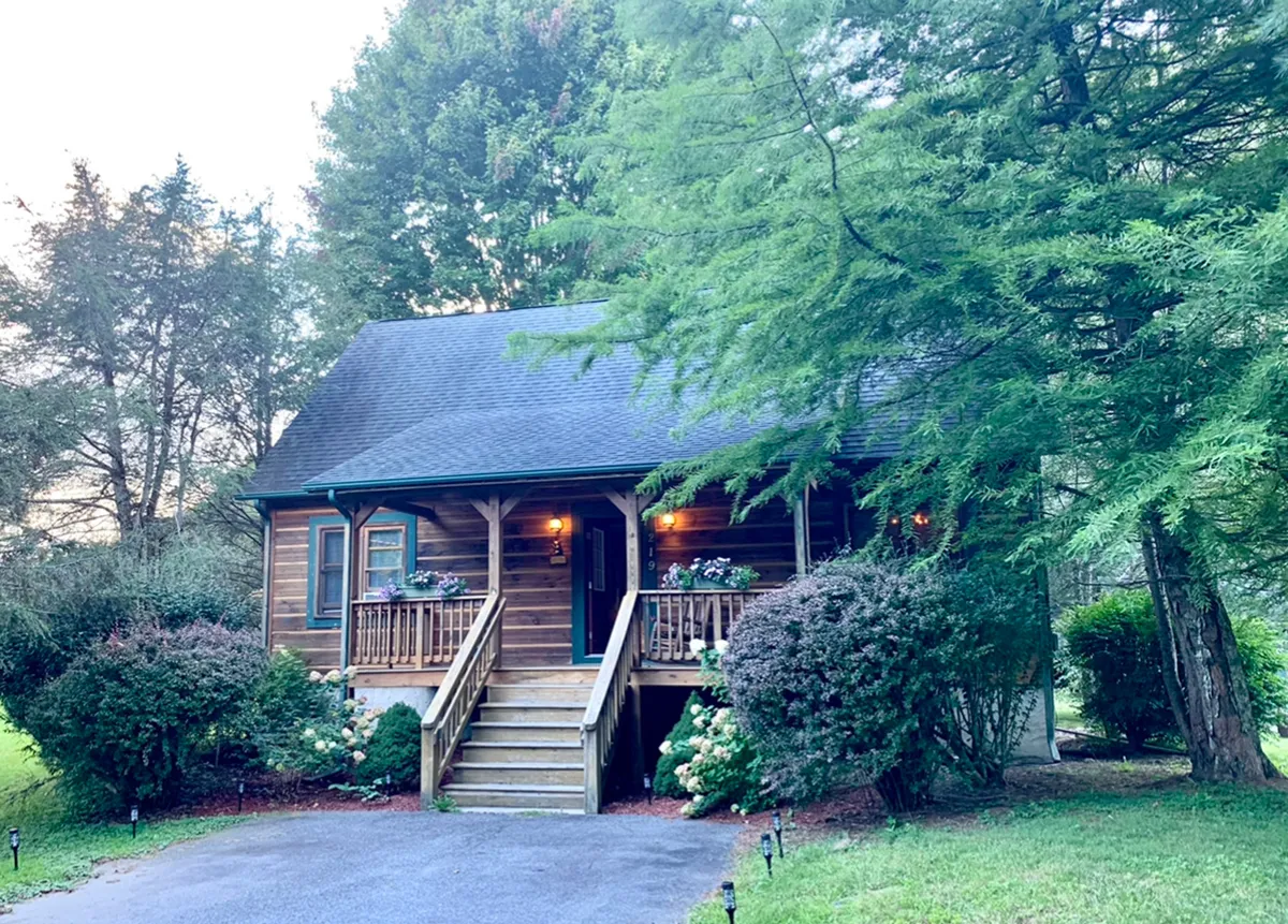 The image shows the front exterior of a charming wooden cabin nestled among lush greenery, with a warmly lit front door and an inviting porch perfect for relaxing. The broad steps lead up to the porch, which is surrounded by trees, creating a peaceful, secluded atmosphere ideal for unwinding or socializing outdoors.