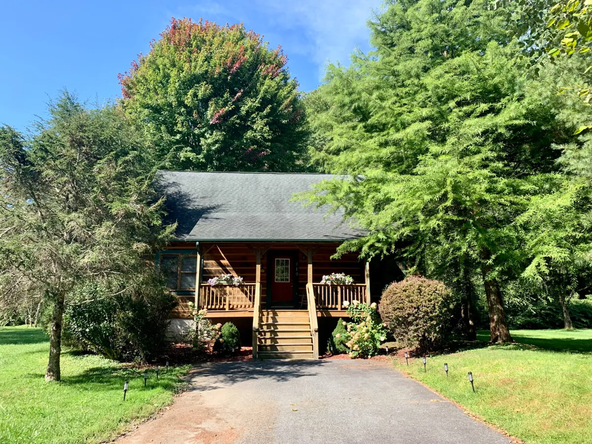 The image features the welcoming front porch of the cabin framed by vibrant greenery. The wooden structure and seating on the porch highlight opportunities for outdoor relaxation and taking in the natural setting after a day of exploring Boone.