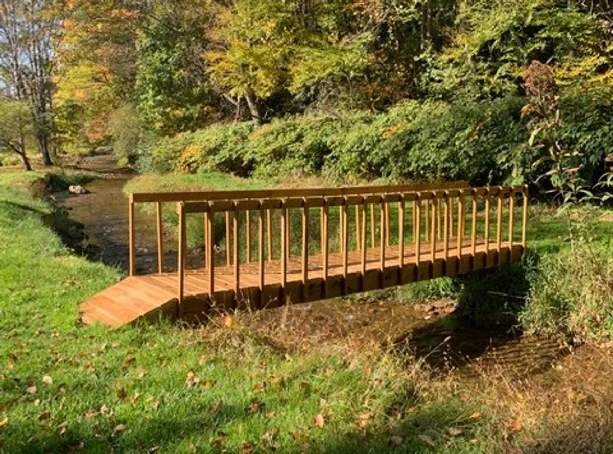A picturesque wooden footbridge spans a clear, flowing stream, surrounded by lush greenery. Although the section notes that the ornamental bridge is under restoration, the image still evokes a sense of exploring creekside paths and appreciating the property's natural beauty, with opportunities for wildlife spotting and peaceful walks.