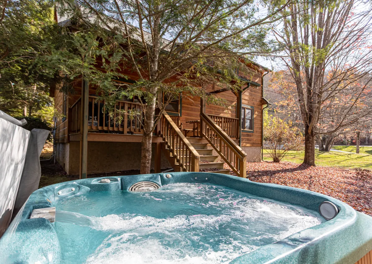 A tranquil exterior view of the wooden cabin nestled among trees, with a prominent hot tub on the deck and a wooden staircase leading up. The setting exudes relaxation and invites guests to unwind after active outdoor pursuits.