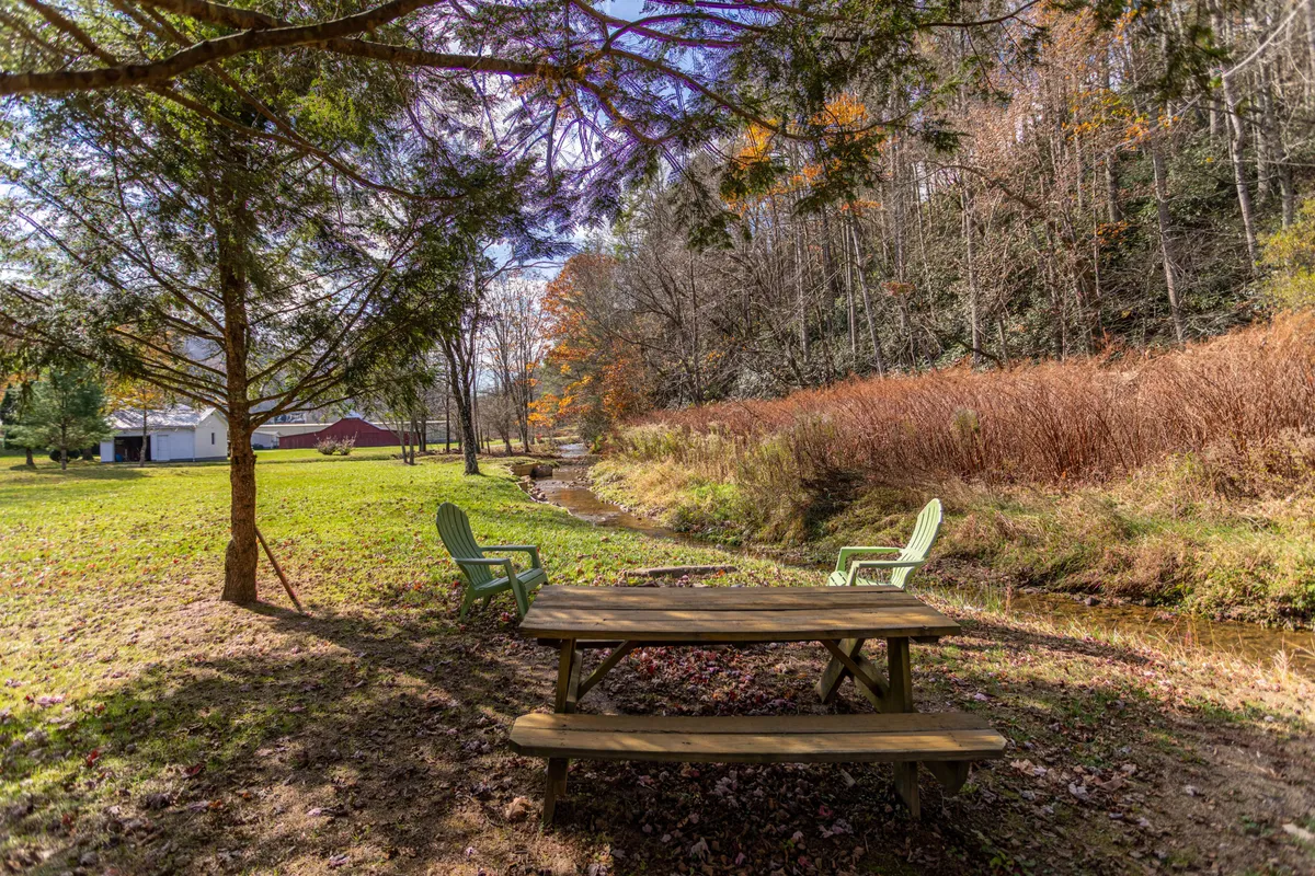 A peaceful backyard setting featuring a picnic table and green chairs nestled beside a gently flowing stream. The area is surrounded by mature trees, creating a tranquil atmosphere ideal for outdoor family activities, picnics, or quiet nature moments. The open lawn space is visible, providing plenty of room for children and pets to play.