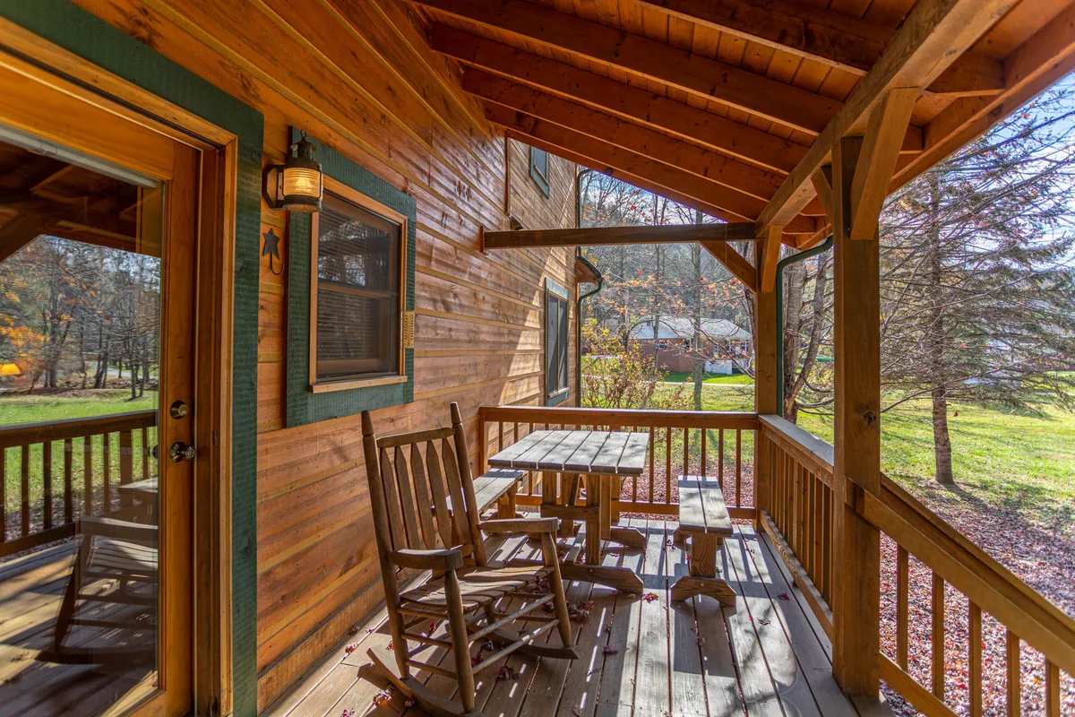 The image features a cozy front porch with wooden rocking chairs and a small outdoor table, set against the backdrop of the cabin's wooden railing. The inviting seating is ideal for sipping coffee in the morning or relaxing in the evening, capturing the spirit of mountain leisure.