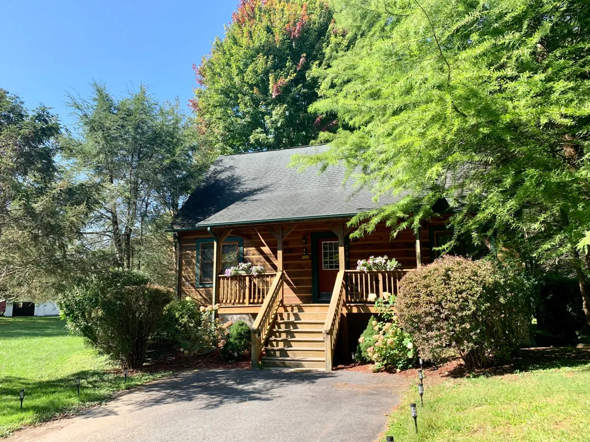 A charming log cabin exterior with a welcoming wooden staircase, flower pots on the porch, and lush greenery surrounding the home. The covered porch invites relaxation and offers a seamless connection to the natural setting.