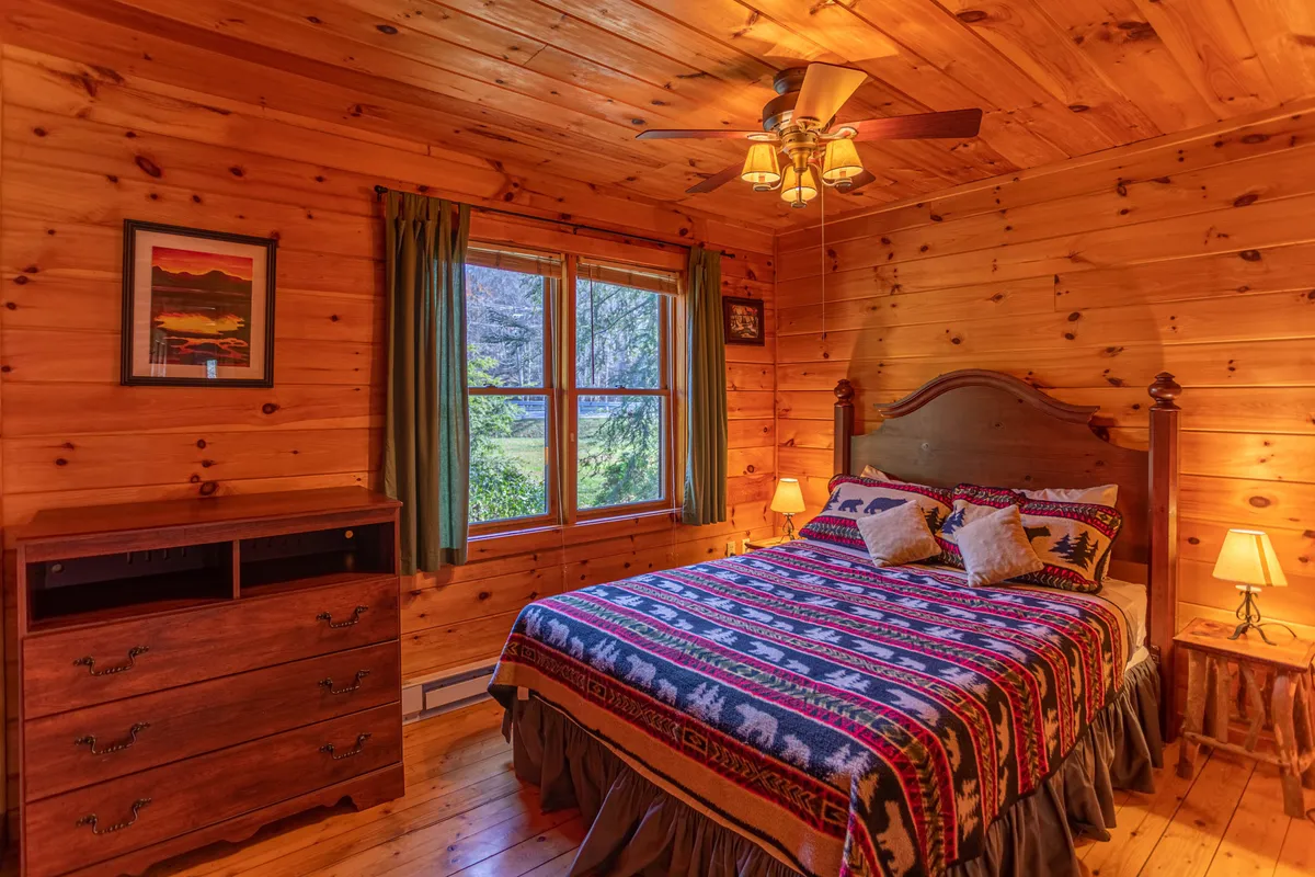 A bedroom featuring a bed with patterned bedding, a window that frames a peaceful outside view, a wooden dresser, and a nightstand with a lamp. The space looks both practical and cozy, perfect for relaxing nights and mornings.