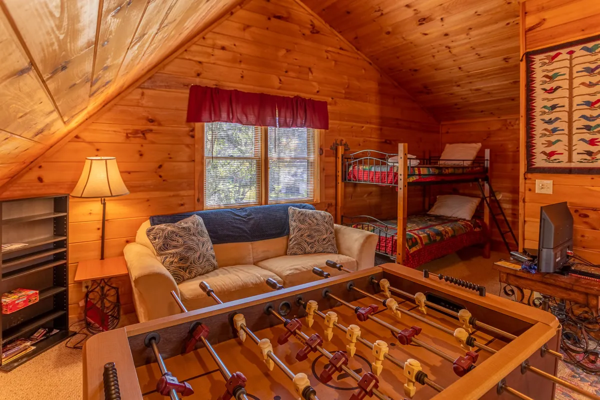 This image showcases the loft's central features: a foosball table in the foreground, a cozy sofa, and bunk beds in the background, all set against warm wooden walls and soft lighting. The space feels playful and relaxed, clearly inviting both games and downtime. The overall arrangement highlights both the recreational and sleeping aspects of the loft.