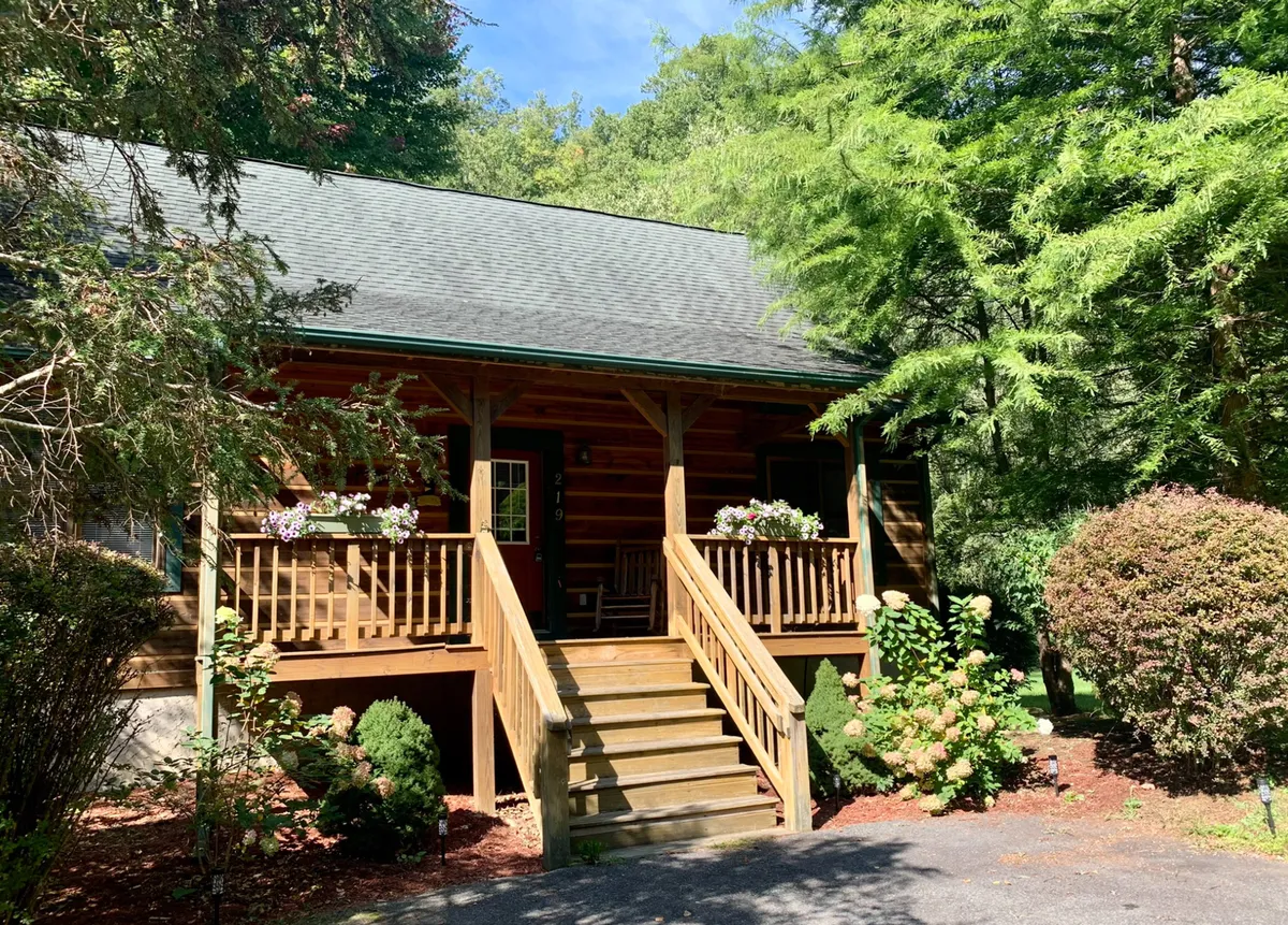 A wide view of the log cabin’s exterior highlighting wooden steps, flower pots, the porch, and surrounding greenery. The perspective invites the viewer to imagine arriving home after a day exploring, ready to relax on the deck or porch.
