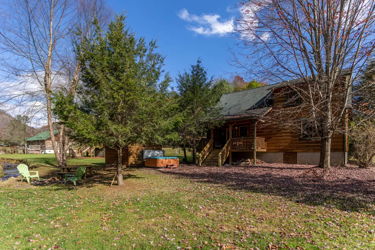 This exterior image showcases the log cabin structure nestled among mature trees, with a view of the outdoor seating area and a hot tub. The tranquil setting is complemented by the lush greenery, creating an inviting atmosphere for relaxation and gathering.