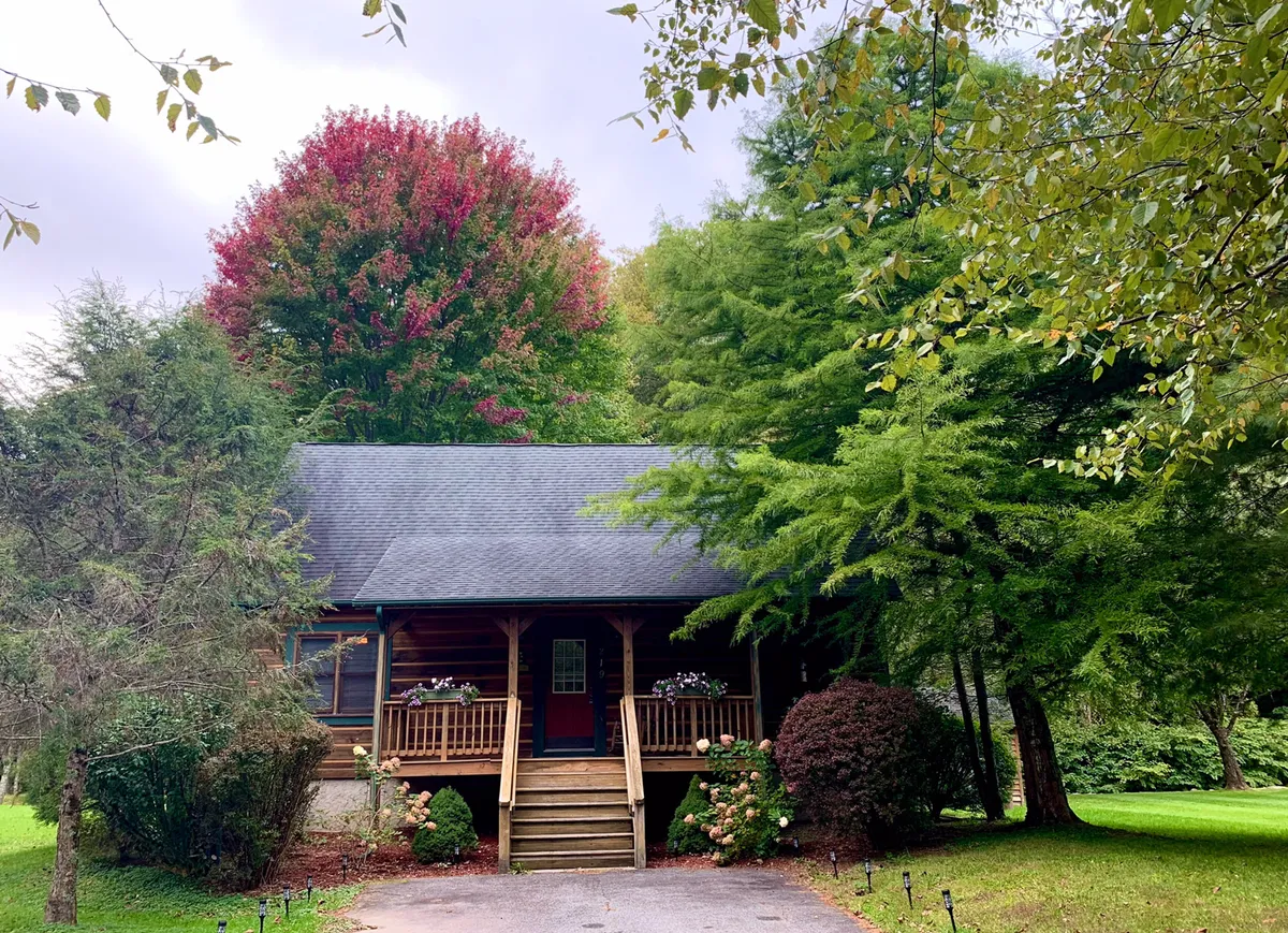 A welcoming wooden porch adorned with colorful autumn trees and classic front steps leading into the cabin. The porch invites relaxation with ample seating and a scenic view of the yard and trees beyond, evoking a sense of warmth and togetherness in a natural setting.