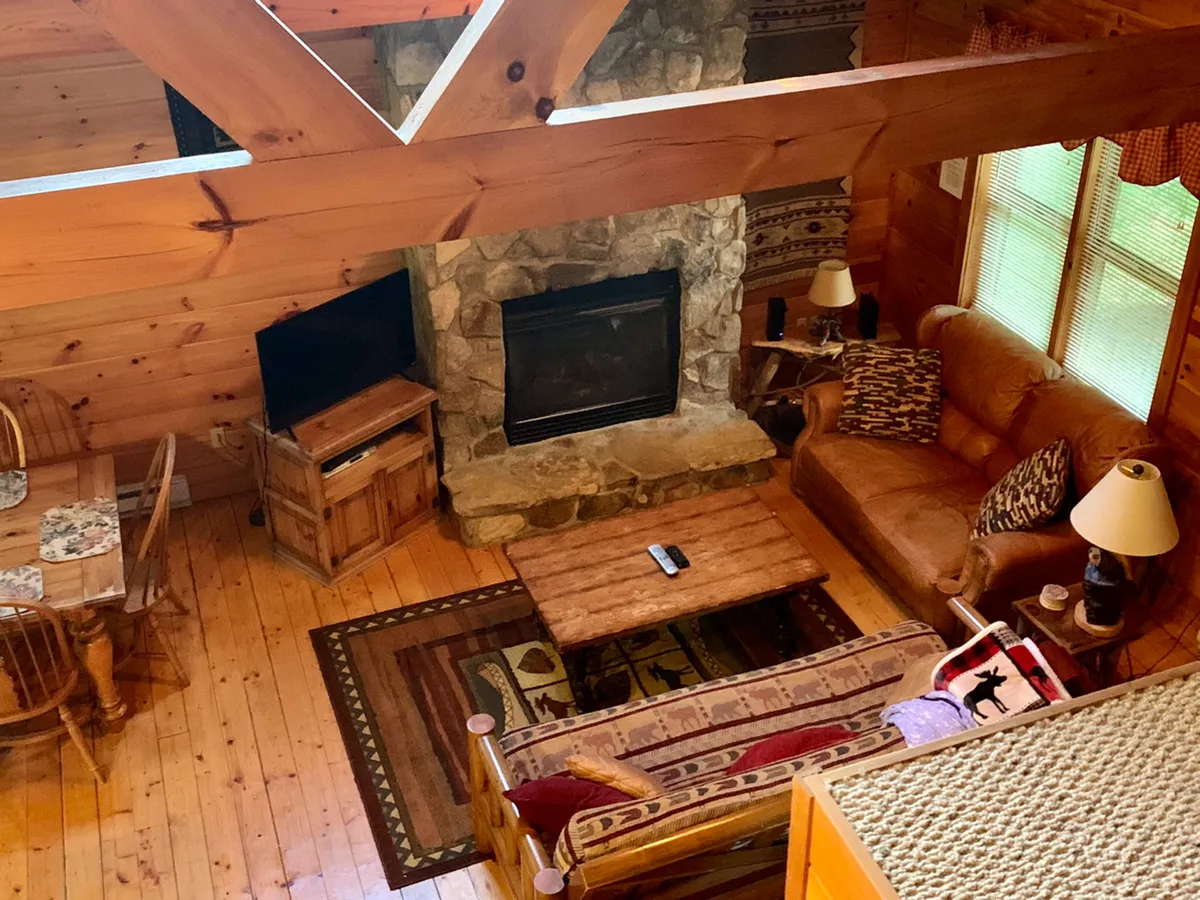 A view of the open-concept living and dining area, featuring a rustic wooden dining table near a stone fireplace, with a comfortable seating area and decorative throw pillows. The space is styled for both family gatherings and relaxed evenings, with a warm and welcoming ambiance.